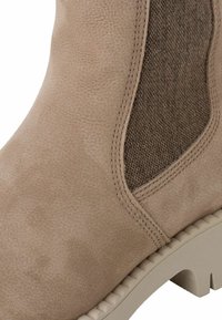 Beige enkelboot met een suede afwerking en een getextureerd tan zijpaneel, voorzien van een lichtgekleurde rubberen zool en zichtbare stiksels.
