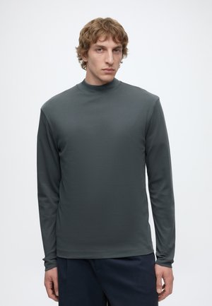 Junger Mann mit hellbraunen lockigen Haaren trägt ein dunkelgraues Langarmshirt und marineblaue Hose und steht vor einem schlichten hellen Hintergrund.