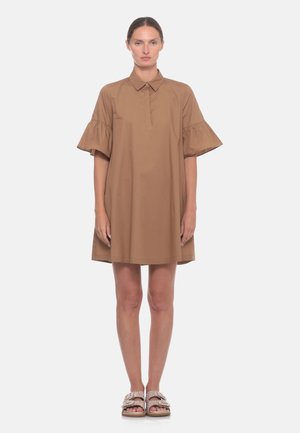 Donna in piedi che indossa un vestito marrone largo con maniche a sbuffo a metà e sandali beige senza lacci su uno sfondo chiaro e uniforme.