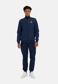 Chaqueta de chándal azul marino con cremallera y cuello alto, acompañada de pantalones jogger a juego. Ambos tienen una textura suave y un diseño ajustado. Zapatillas blancas.