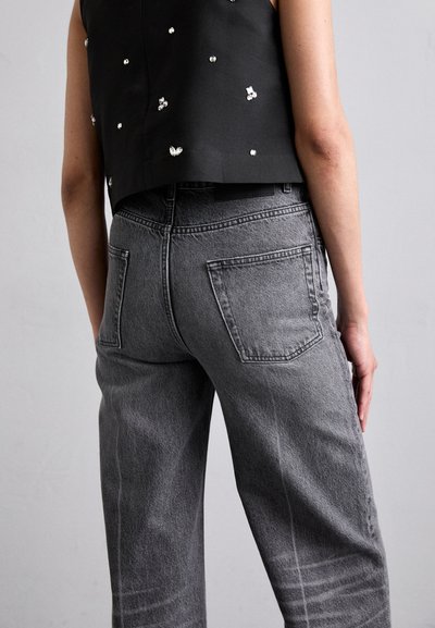 Črn krojeni top z srebrnimi okrasnimi elementi, v kombinaciji z visokimi sivo-nazaj zaključenimi kavbojkami s pravimi hlačnicami, ki imajo zadnje žepe in zbledelo teksturo.