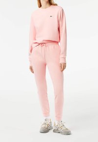 Sudadera rosa y joggers a juego en tejido suave, con cuello redondo, cinturilla elástica y un pequeño logo verde. El calzado son zapatillas grises.