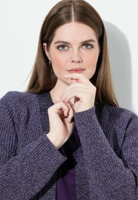 Kobieta o długich brązowych włosach ubrana w fioletowy dziergany kardigan i koszulę, trzyma ręce blisko twarzy na jednolitym jasnym tle.