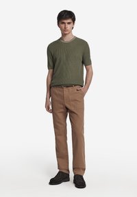 Olivgrön, texturerad kortärmad tröja i stickad kvalitet kombinerad med ljusbruna, löst sittande byxor. Svarta skor fulländar outfiten.