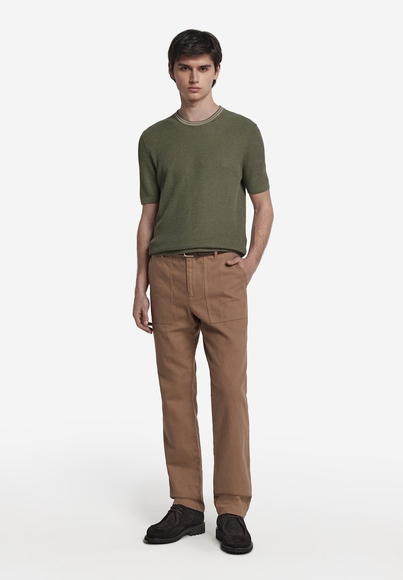 Olivgrön, texturerad kortärmad tröja i stickad kvalitet kombinerad med ljusbruna, löst sittande byxor. Svarta skor fulländar outfiten.