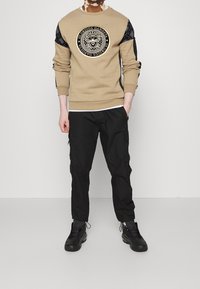 Sudadera beige con un logo negro redondo en el centro y mangas negras con patrón, combinada con pantalones cargo negros y zapatillas negras.
