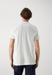 Hombre con cabello castaño corto, vistiendo una camiseta blanca de manga corta y jeans oscuros, de pie frente a un fondo gris claro.