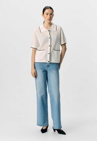 Kortärmad, vit prickig blus med svart kant och två framfickor, i kombination med ljusblå vida jeans och svarta spetsiga skor.