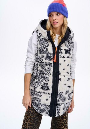 Žena v bílé mikině s kapucí, černo-bílém vzorovaném fleecovém vestě s kapucí, kalhotách s leopardím vzorem a pletené čepici modro-červené barvy.