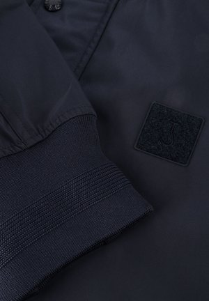 Navy blue jacket featuring a smooth exterior, textured black cuff, and square felt patch with a logo. Buttons are visible on the front.