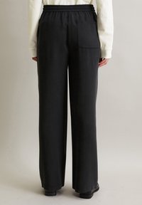 Personne portant un pantalon noir ample avec une taille élastique et une poche arrière, associé à un haut à manches longues blanc et des chaussures noires, debout.