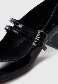 Black patent leather Mary Jane shoe featuring double thin straps with silver buckles and a block heel on a white background.