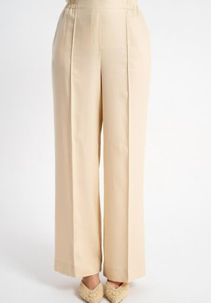 Beżowe, wysokie w talii spodnie z szerokimi nogawkami i przeszyciami z przodu, noszone z beżowymi plecionymi balerinami z ostrym noskiem na białym tle.