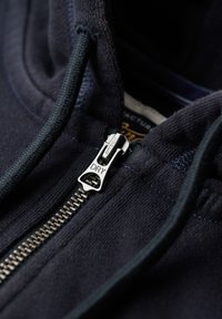 Marineblå lynlås hoodie med en sølvmetal lynlås med "DRY" tekst, tekstureret stof og kontrastsyninger omkring hætteåbningen.