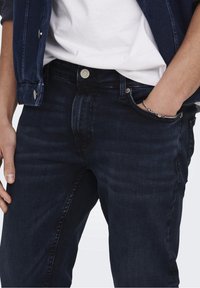 Mörkblå denimjeans med en något sliten struktur. Har en klassisk femficksdesign och en silverfärgad knäppning i midjan.