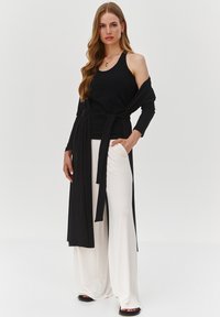 Czarna top bez rękawów warstwowany z czarnym okryciem, mający długie rękawy i wiązanie w talii, w połączeniu z białymi szerokimi spodniami i czarnymi sandałami.