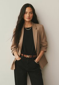 Blazer color canela sobre una camisa negra, combinado con pantalones negros. Cinturón marrón con una hebilla dorada ornamentada. Cabello largo y oscuro, ligeramente ondulado.