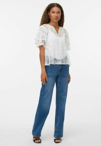 Haut en dentelle blanche avec de courtes manches voler, un décolleté en V, design superposé, porté avec un jean bleu taille haute et des sandales noires à brides.