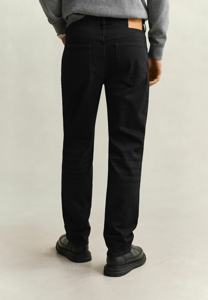Homme portant un jean noir coupe droite, des chaussures noires et un haut gris à manches longues, debout sur un fond clair et uni.