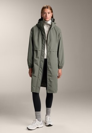 Lange olijfgroene gewatteerde jas met een capuchon, elastische manchetten en twee voorzakken. Draagt over een lichtgrijze ritstrui en leggings.