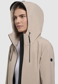 Ljust beige huvjacka med svart dragkedja och justerbara snoddar, med en elegant design och texturerad yta.