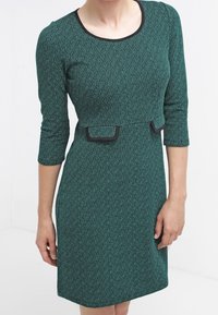 Vihreä teksturoitu mekko, jossa on kolme neljänneshihaa, pyöreä kaula-aukko ja mustat yksityiskohdat vyötäröllä. Slim fit ja polvipituinen muotoilu.