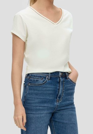 Femme portant un haut blanc uni à manches courtes avec col en V, rentré dans un jean bleu taille haute, une main dans la poche, sur un fond clair.