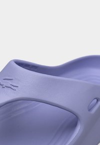 Gros plan sur une sandale en caoutchouc mauve clair avec une bride ornée d'un logo crocodile en relief.