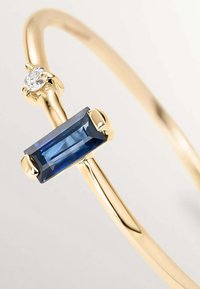 Pulsera de oro con una piedra preciosa azul rectangular y una piedra clara redonda, con un acabado suave y pulido y un diseño minimalista.