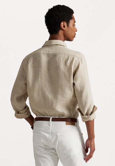 Chemise en lin beige avec col et manches retroussées, portée avec un jean blanc et une ceinture en cuir marron, montrant un tissage texturé.