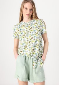 Top de flores de mangas curtas com fundo branco, apresentando flores azuis, verdes e castanhas. Combinado com calções verdes claros com um detalhe de laço.