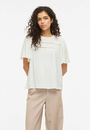 Femme aux cheveux bouclés portant un chemisier blanc ample avec des détails en dentelle et un pantalon beige à jambes larges, debout devant un fond blanc.