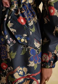 Jachetă din material întunecat cu imprimeu floral, buzunar cu capse, manșetă elastică și logoul vizibil „Joules” lângă buzunar, purtată de o persoană cu inele.