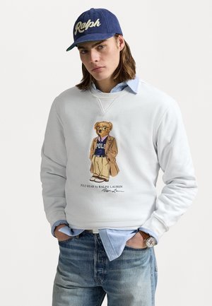 Sudadera blanca con una ilustración de un oso en traje formal. Combinada con una gorra azul marino, una camisa azul clara y vaqueros. Accesorios destacados incluyen un reloj.