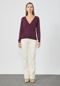 Burgunderfarbener Cardigan mit tiefem V-Ausschnitt, vorderem Knopfverschluss, langen Ärmeln und gerippten Bündchen, kombiniert mit cremefarbenen Schlaghosen und beigen Absatzschuhen.
