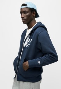 Μπλε ζακέτα με φερμουάρ σε ναυτικό μπλε χρώμα με μεγάλο λευκό λογότυπο στο στήθος. Διαθέτει τσέπη καγκουρό, ριμπ μανσέτες και λογότυπο της Nike στο μανίκι.