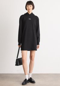 Čierne šaty s kapucňou a dlhými rukávmi, s klokanou kapsou a malým logom na hrudi. Model drží čiernu tašku, má na sebe biele ponožky a mokasíny.