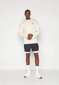 Krémová mikina s malou nášivkou, spárovaná s tmavými šortkami s bílým lemem. Model v bílých teniskách stojí na neutrálním pozadí.