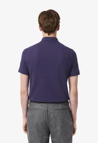 Lila kortärmad polo med krage, sedd bakifrån, ihopparad med grå strukturerade byxor. Minimal detaljering, klassisk passform.