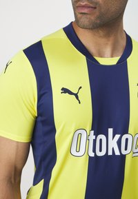 Puma-Sporttrikot mit gelben und marineblauen vertikalen Streifen. Mit Rundhalsausschnitt, kurzen Ärmeln und dem "Otokog"-Logo auf der Vorderseite.