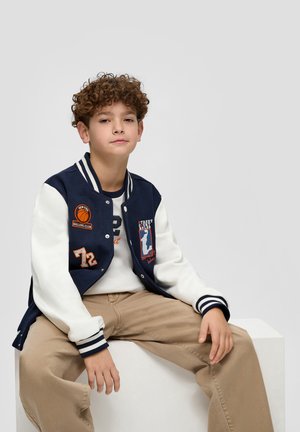 Marineblau- und weiße Varsity-Jacke mit gestickten Aufnähern, gestreiften Bündchen und Knopfverschluss, kombiniert mit einem beigen Hemd und khakifarbenen Hosen.