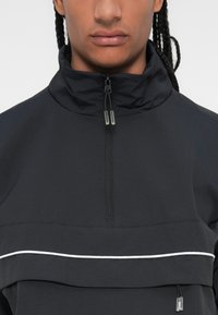 Giacca nera con zip e colletto alto, caratterizzata da una striscia orizzontale bianca e un cordoncino regolabile all'orlo. Tessuto con una texture liscia.