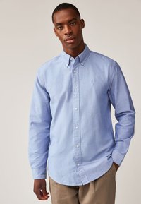 Hellblaues Hemd mit Knopfleiste, Kragen und langen Ärmeln. Verfügt  über eine dezente Textur, vordere Knöpfe und ein kleines Logo auf der Brust.