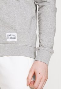 Szary organiczny bawełniany sweter z rękawami zakończonymi ściągaczem, z metką z napisem "NIE PANIKUJ, TO ORGANICZNE" w pobliżu dolnego brzegu.