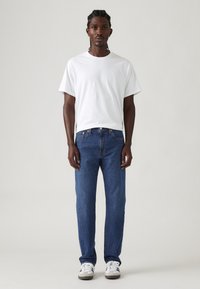 Hombre de pie mirando hacia adelante, vestido con una camiseta blanca sencilla, jeans azules y zapatillas blancas con rayas negras, contra un fondo neutro.