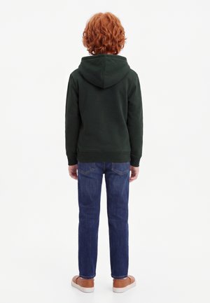 Enfant aux cheveux roux bouclés portant un sweat à capuche vert foncé, un jean bleu et des baskets marron, debout de dos sur un fond uni.
