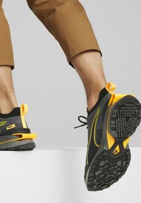 Person, der schwarze und gelbe Puma-Sneaker mit braunen, knöchellangen Hosen trägt, der von einer weißen Fläche tritt und die Schuhsohlen zeigt.