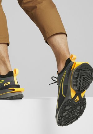 Personne portant des baskets Puma noires et jaunes avec un pantalon court marron, descendant d'une surface blanche montrant les semelles des chaussures.