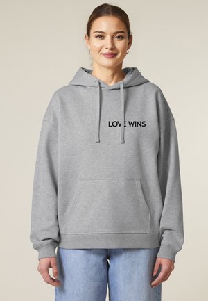 Sweatshirt à capuche gris avec "LOVE WINS" brodé en noir. Il présente une poche avant et des poignets côtelés, associé à un jean bleu clair.
