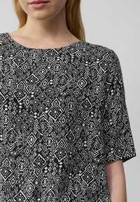 Un tissu à motifs noir et blanc présente des designs géométriques avec des diamants et des carrés. La chemise a un col rond et des manches courtes.